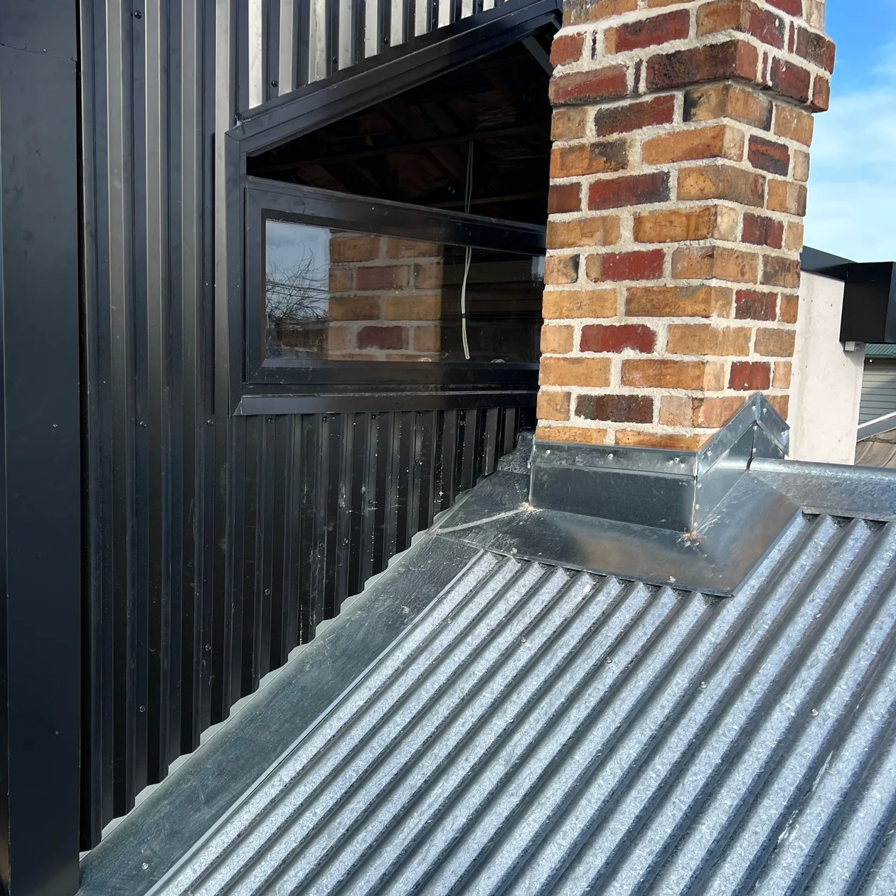Black vertical metal cladding beside a brick chimney, with flashing onto a corrugated roof