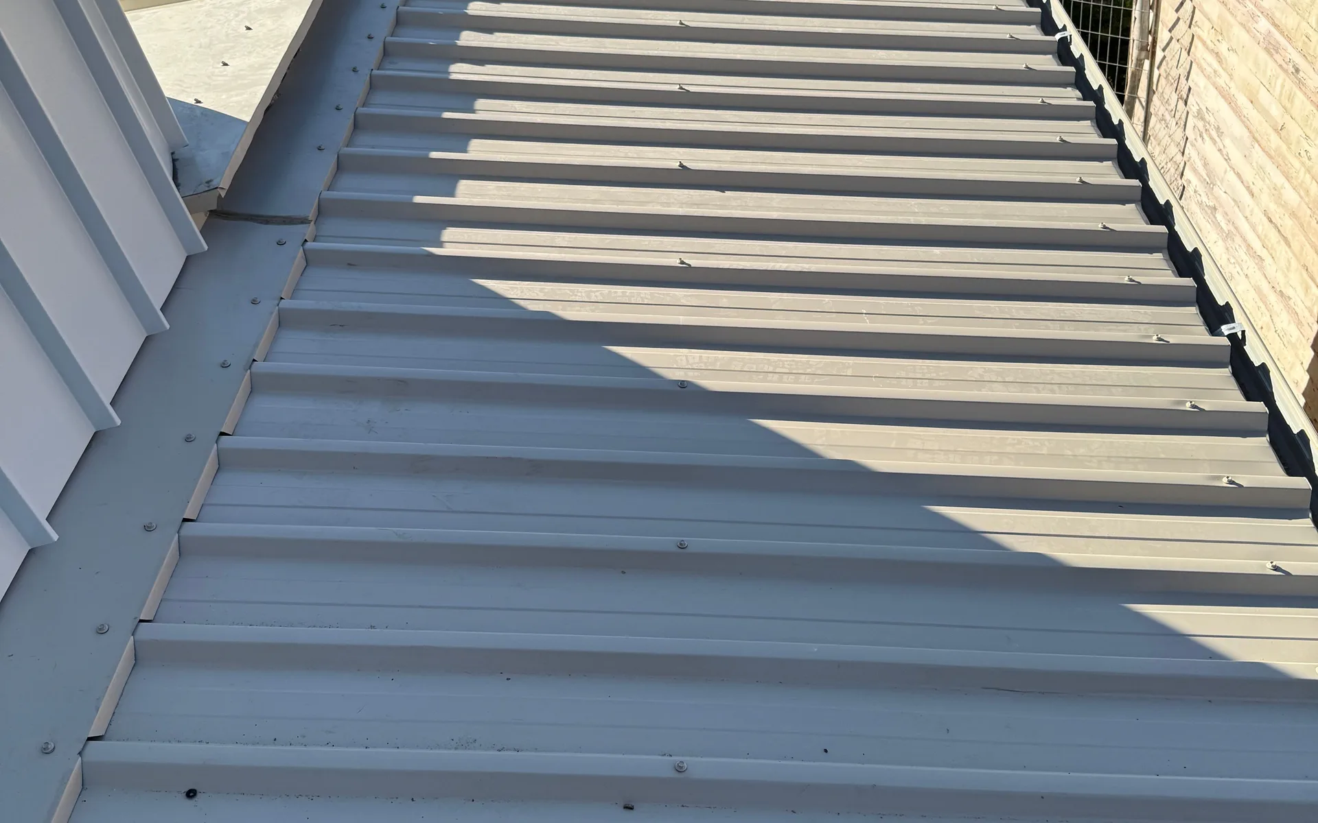 Pale metal roof sheets running beside a flashed wall junction in afternoon sun
