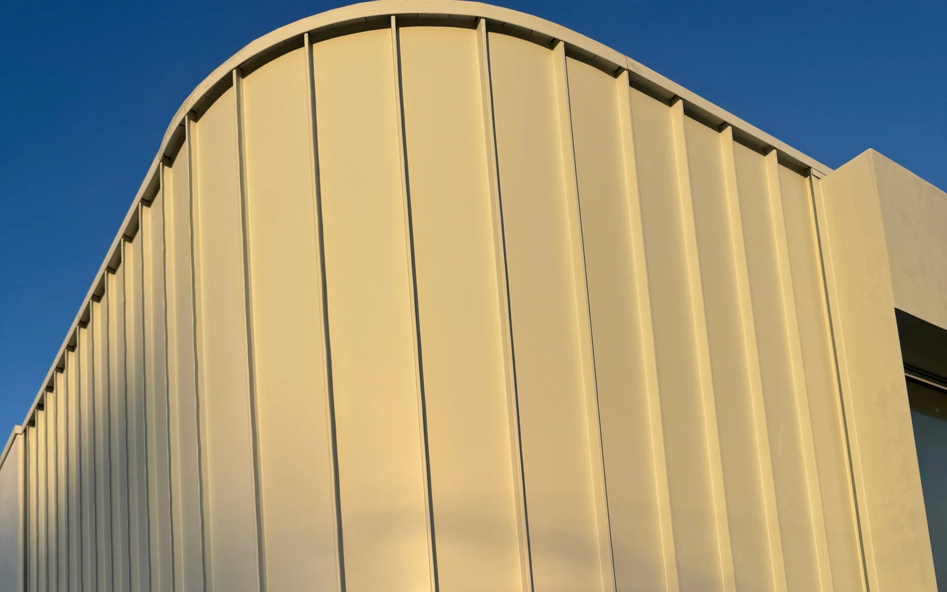 Pale vertical metal cladding curving over the top of a feature wall against a blue sky