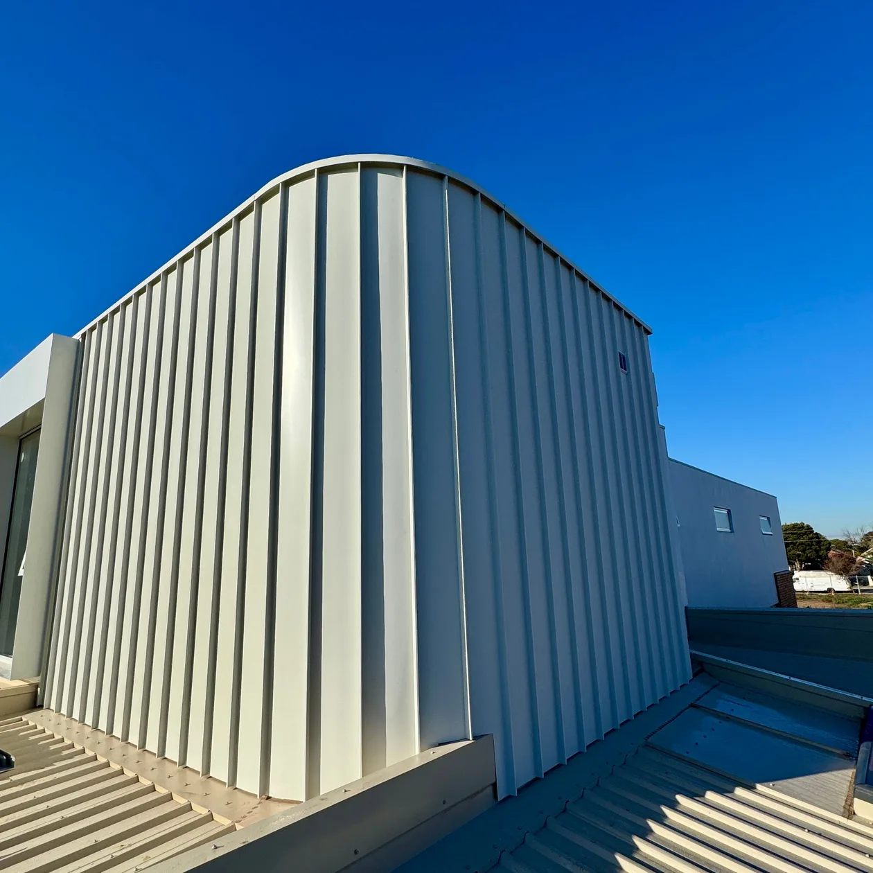 Pale vertical metal cladding curving over the top of an upper-storey wall above a metal roof
