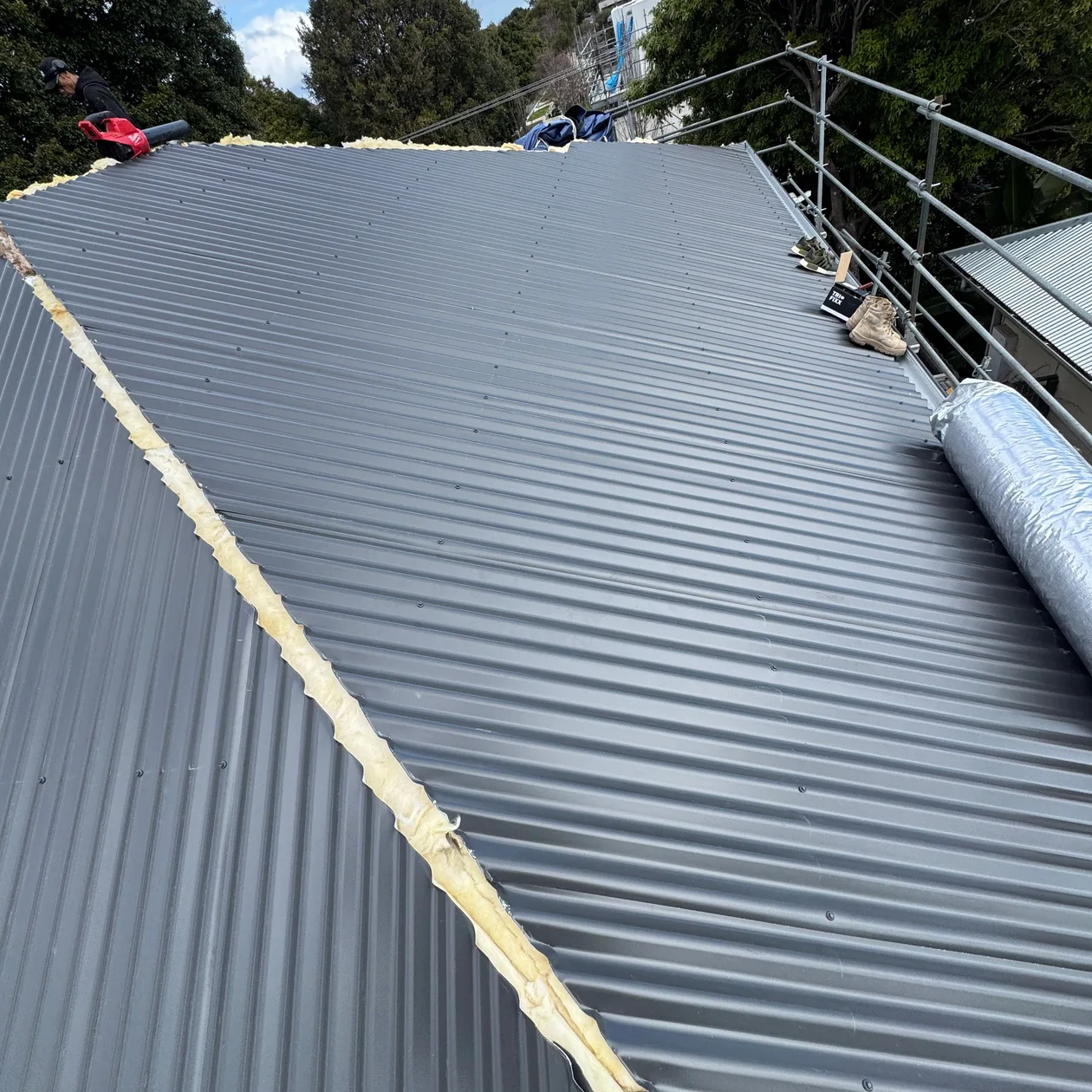 Dark corrugated metal roof sheets partway through installation, with a scaffold rail along the edge