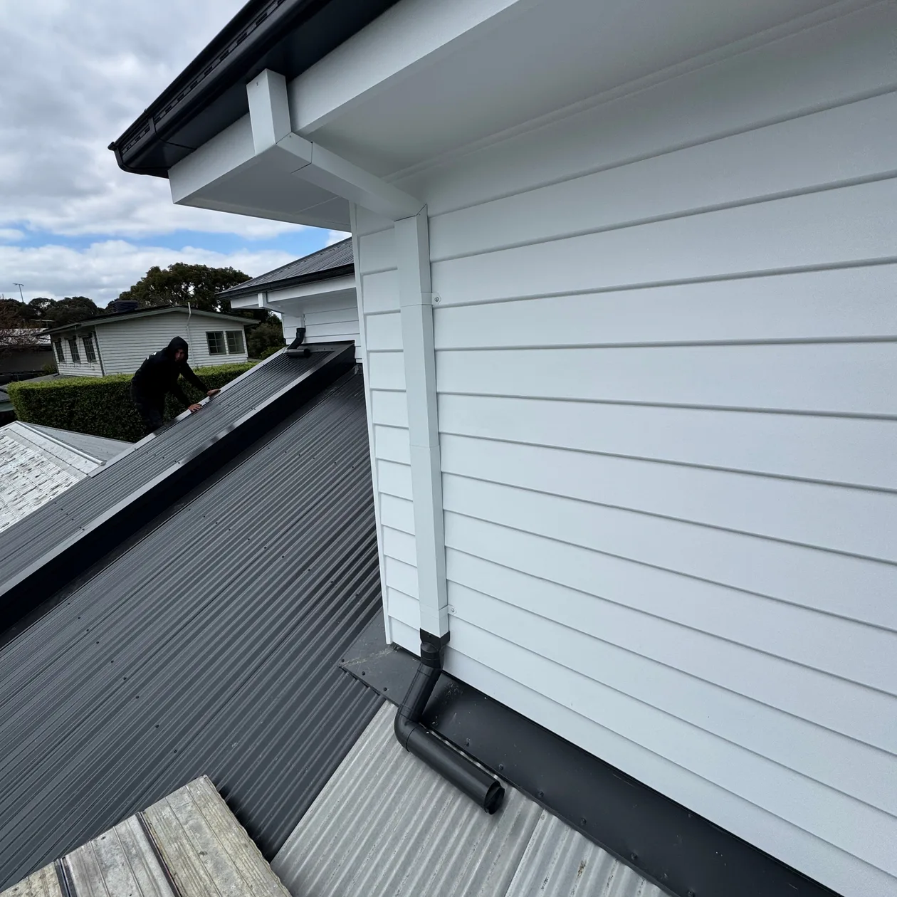 White gutter and downpipe on a weatherboard dormer draining onto the dark metal roof below