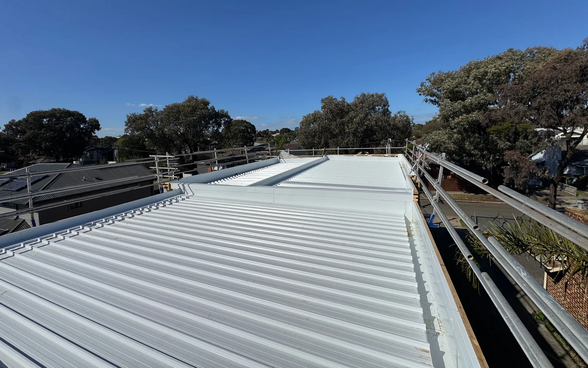 Long pale low-pitch metal roof with an edge rail, trees and rooftops beyond