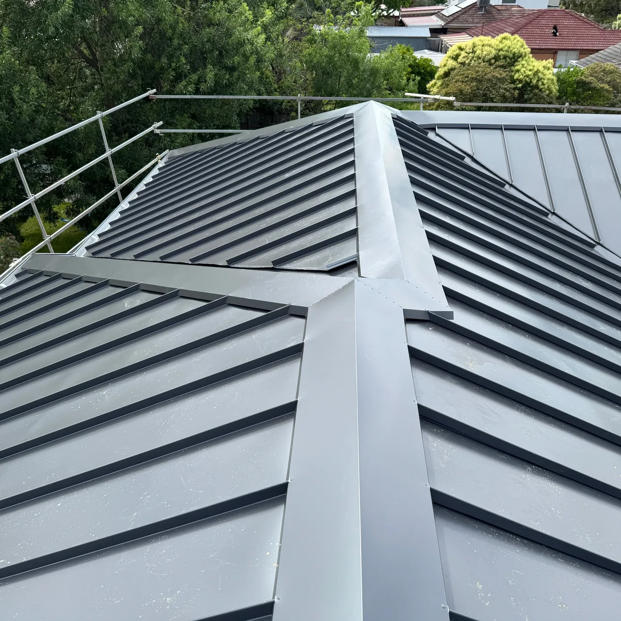 Grey metal hip roof with raised seams and folded hip cappings, trees behind