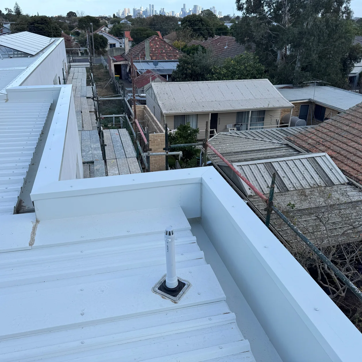 Pale metal roof with a pipe flashing beside a capped parapet wall, rooftops beyond