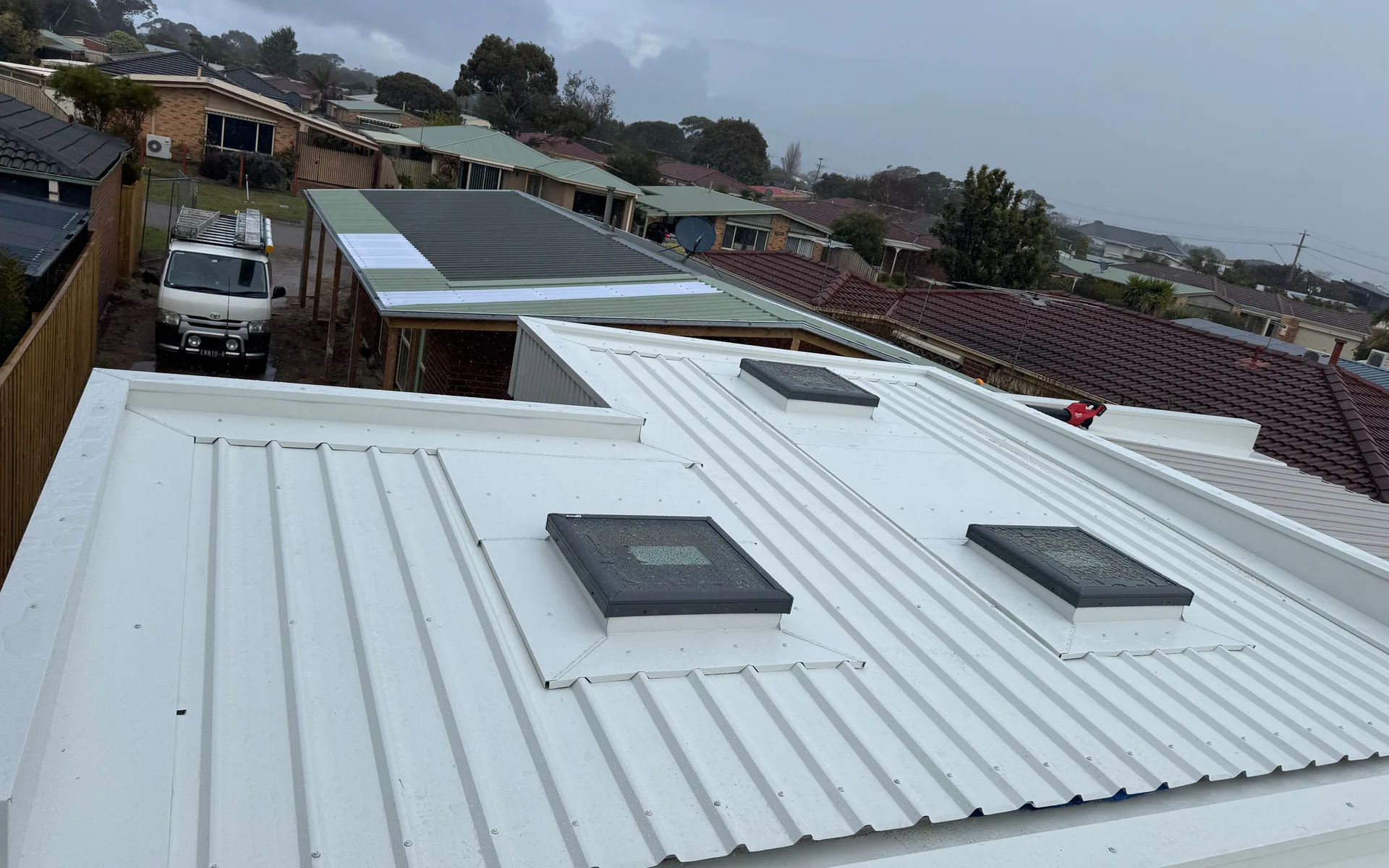 Pale metal roof with two roof windows and suburban rooftops behind